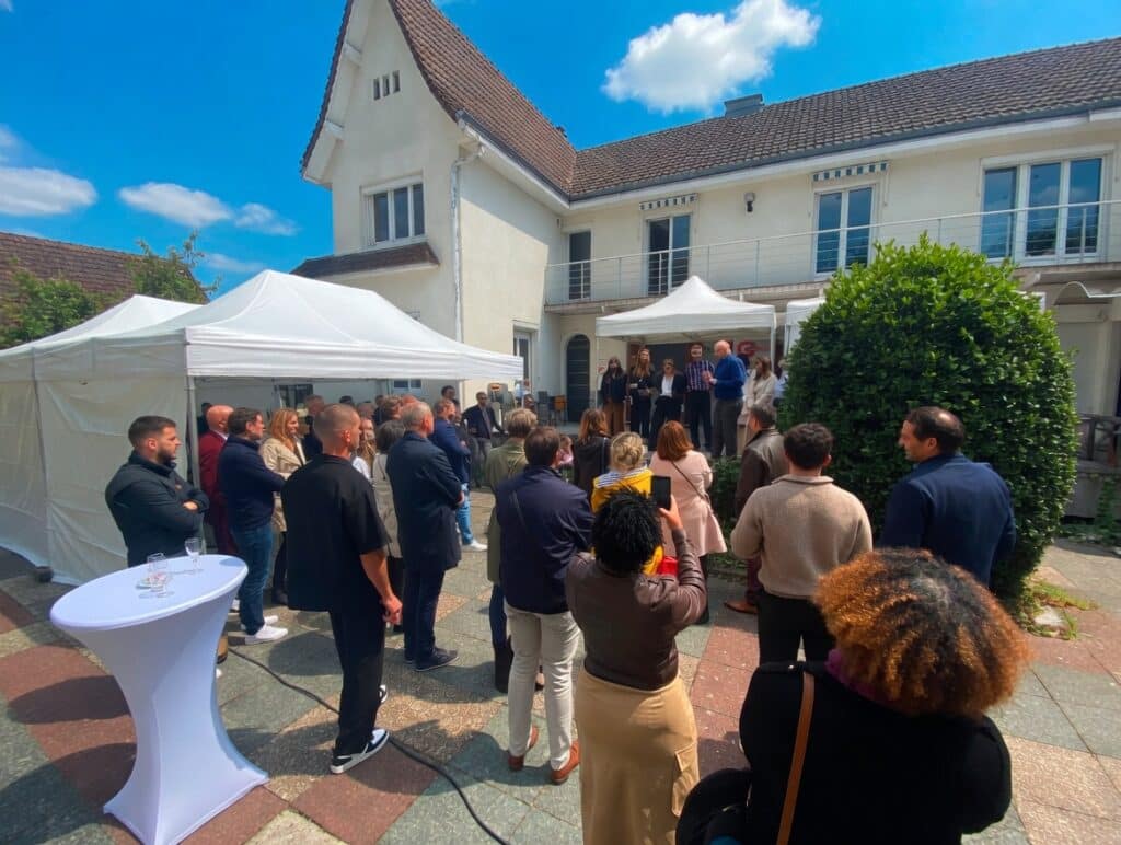 Group of people at an outdoor ceremony in a courtyard, listening to speakers on a small stage under a white tent.
