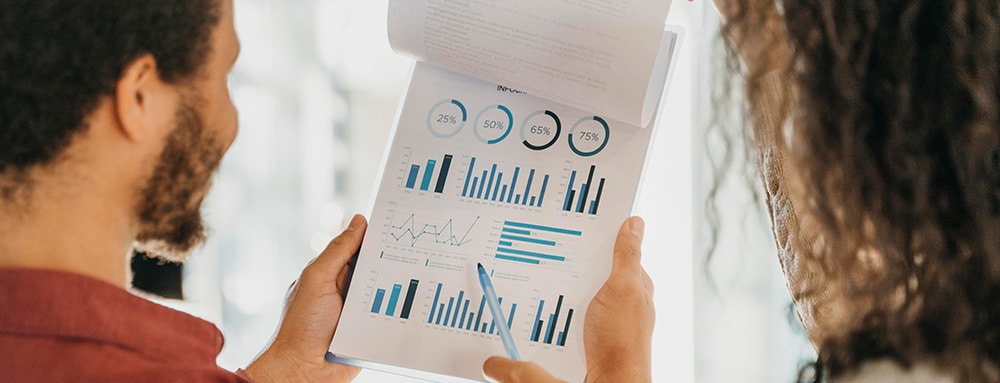 Two people review a clipboard with blue bar charts and pie charts displaying business analytics.
